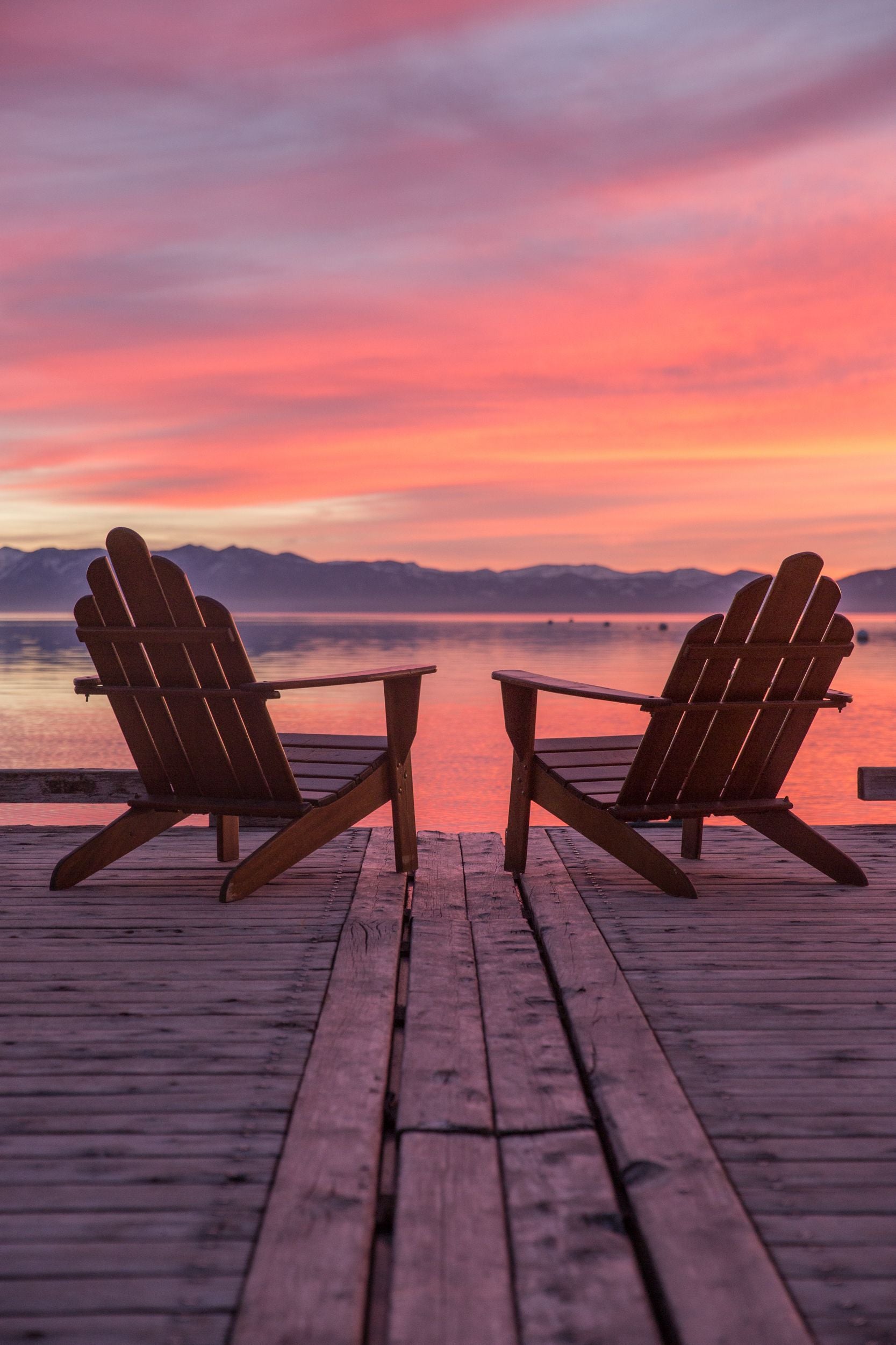 Two Adirondack chairs on a dock overlooking the a multi-color sunset above Lake Tahoe.