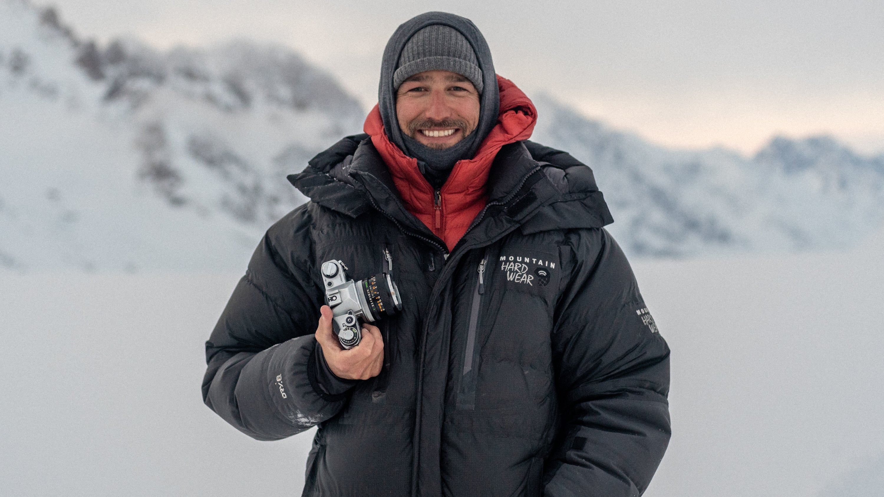 A man bundled up in puffy jackets and a camera in hand stand in front of a snow Alaska mountain range.