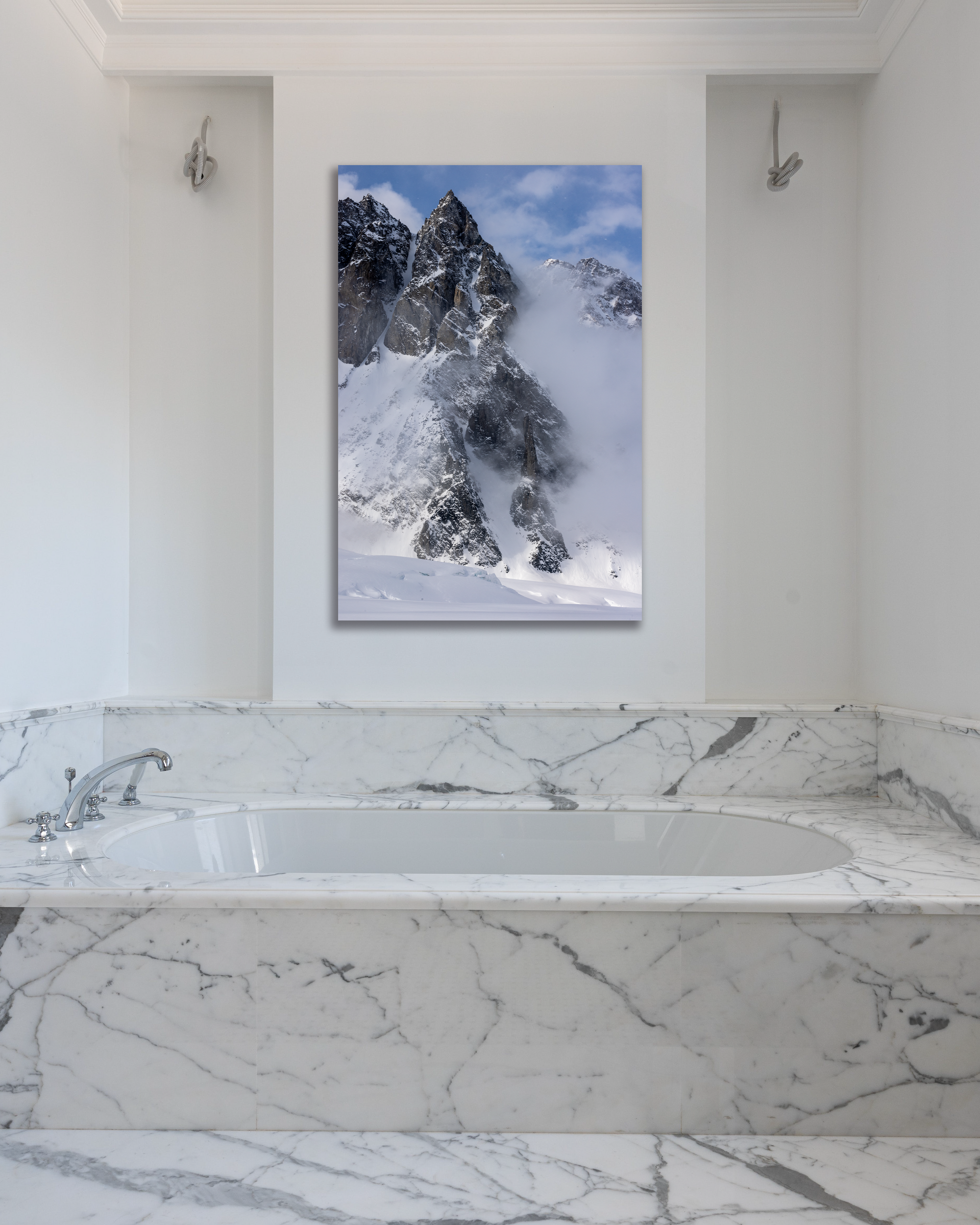 A jagged spire of mountain rocks push through the clouds of a photo hanging above a marbled tub.