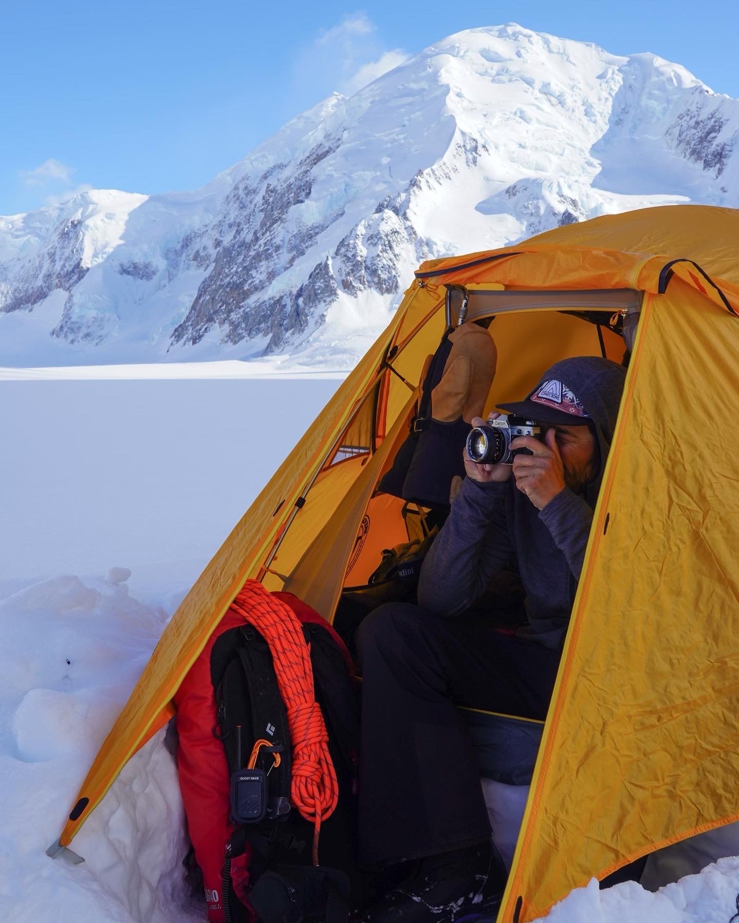 A man sits in the doorway of a yellow tent with a camera to his eye. A snow encrusted mountain peak looms in the background.
