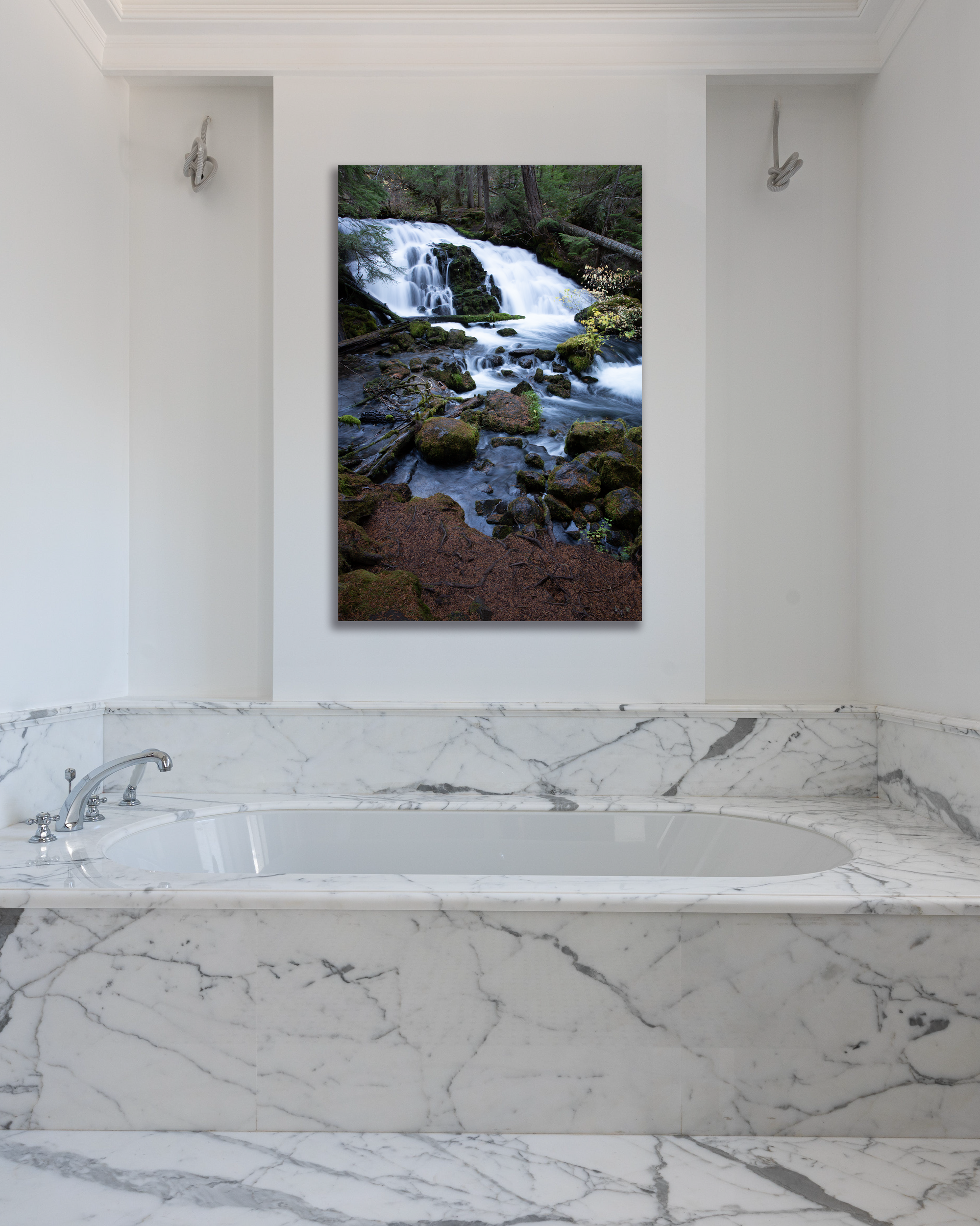 A photograph with a small waterfall surrounded by mossy rocks hangs on a bathroom wall above a bathtub