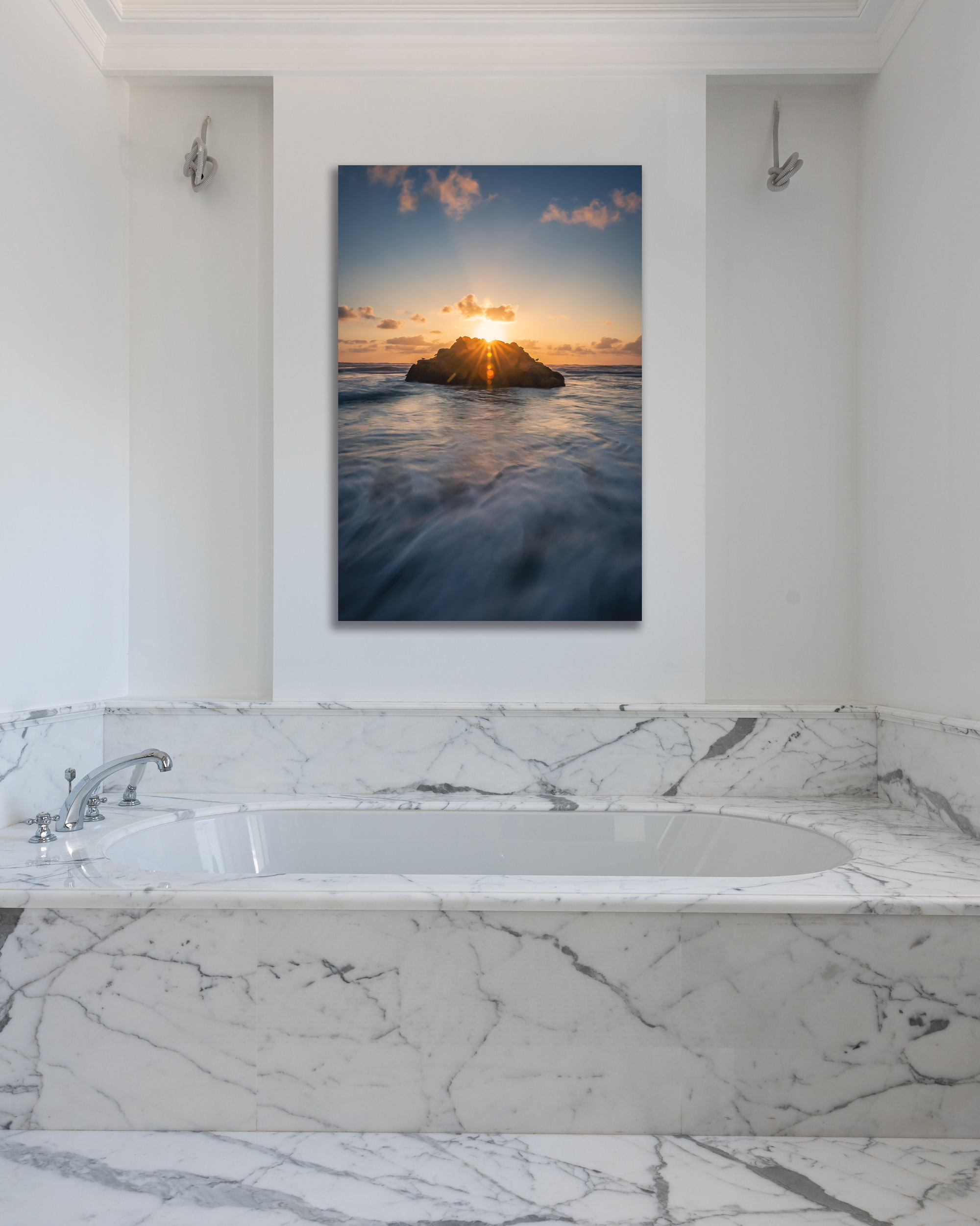 Two rocks enrobed in the coastal waters of the Pacific glow in the sunset in a photograph hung above a marbled bathtub.