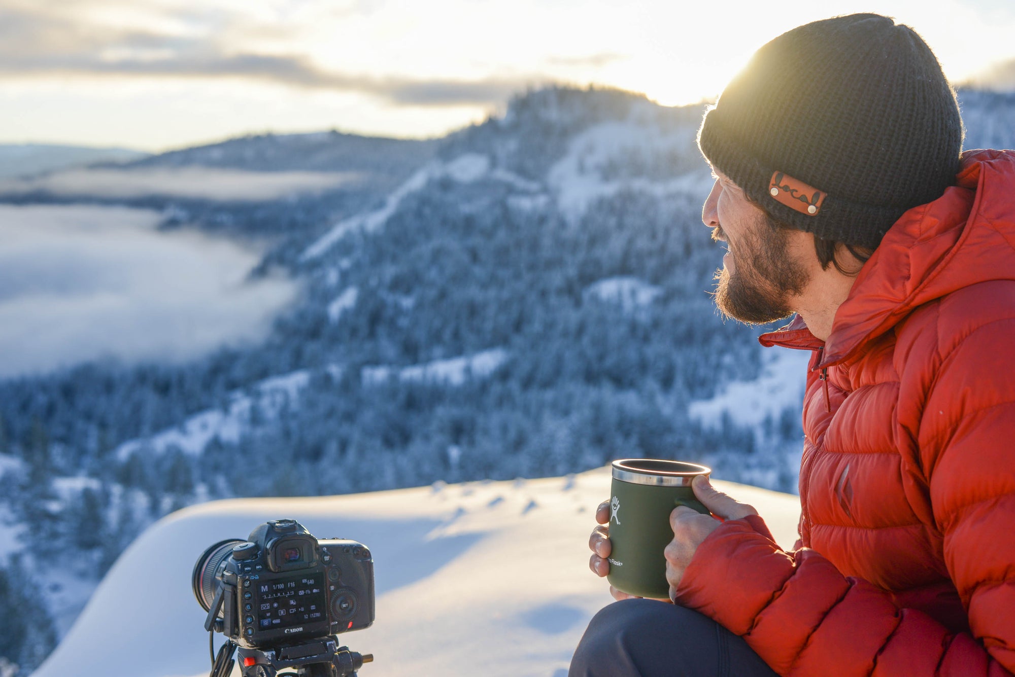 A bearded man wearing a red jacket and black beanie sits above the snowy tree line with a cup of coffee and camera.
