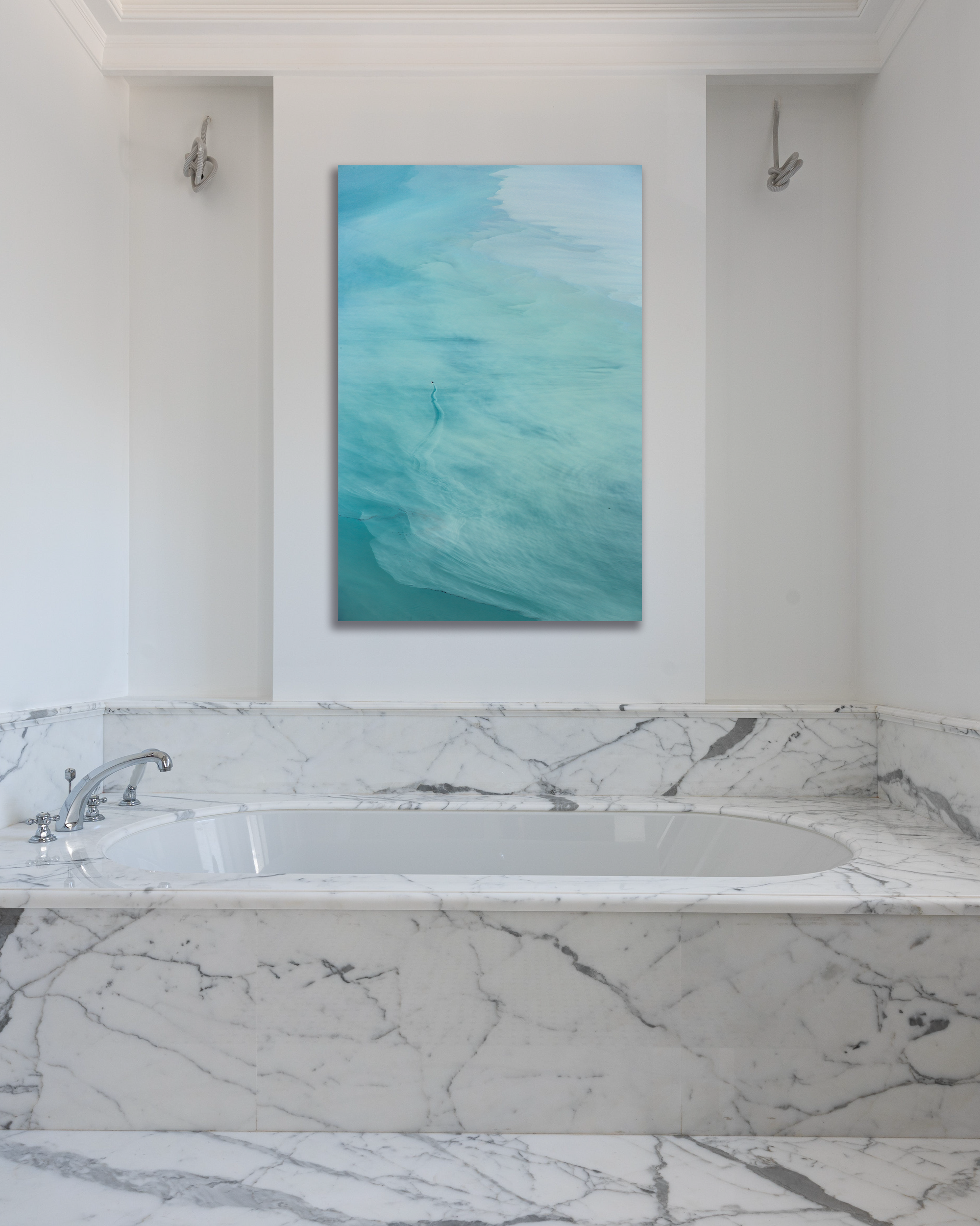 A photograph of turquoise blue waters nearly hide a small boat hangs above a bathtub in a marbled bathroom.