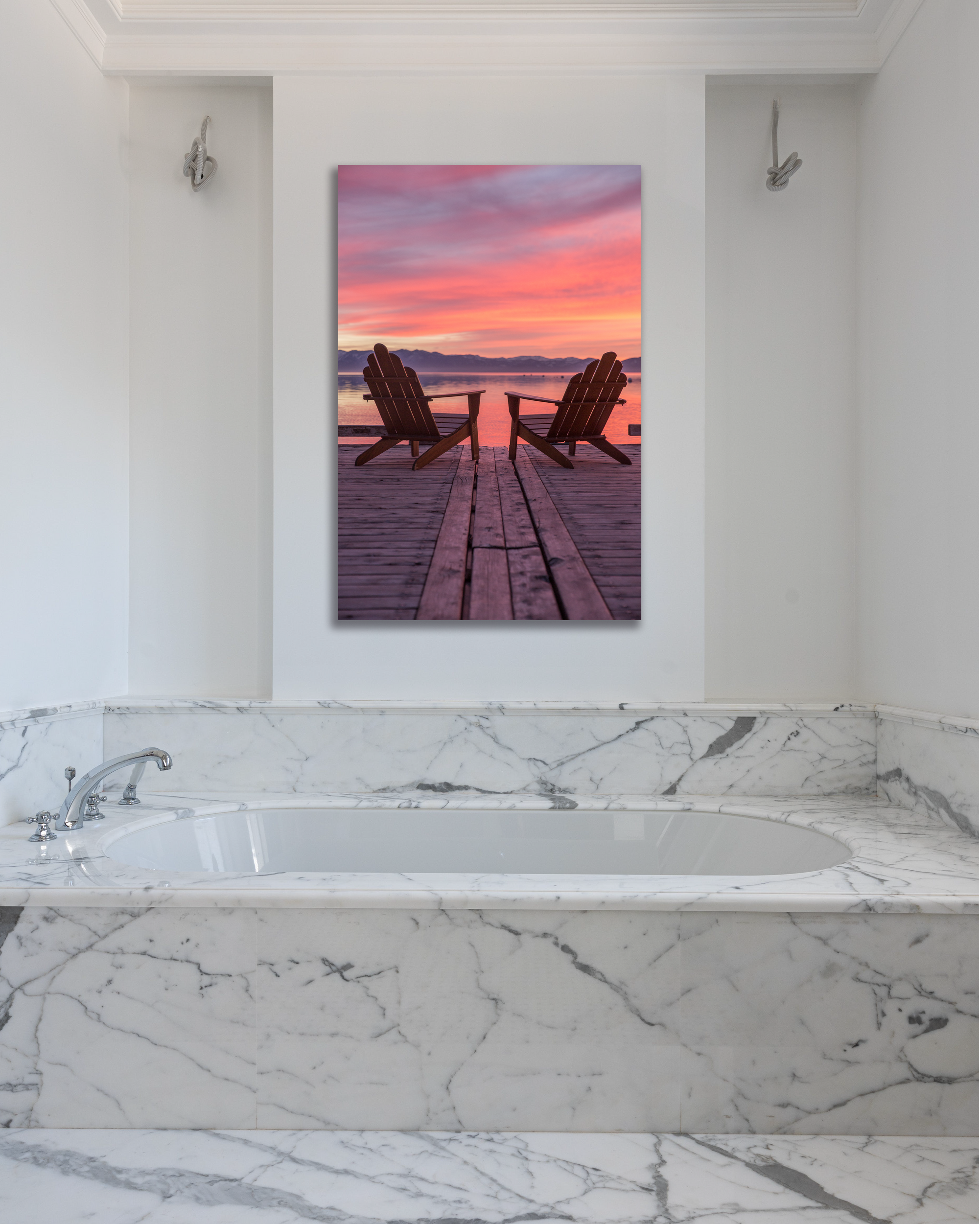 A marble bathroom is decorated with a sunset photograph showing two Adirondack chairs on a lake dock at sunset.