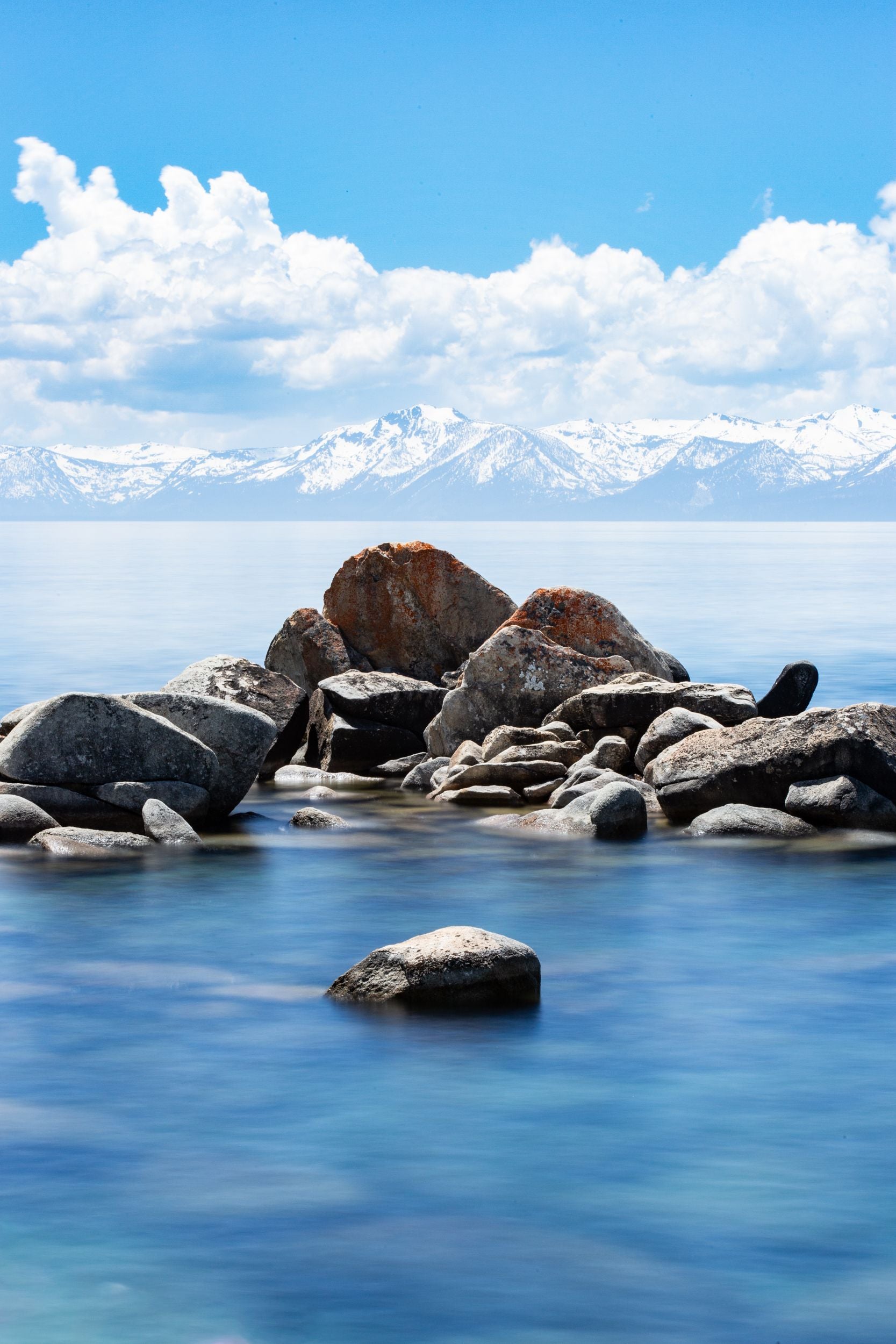 Granite rocks sit in the blue waters of Lake Tahoe as snow capped peaks line the horizon.
