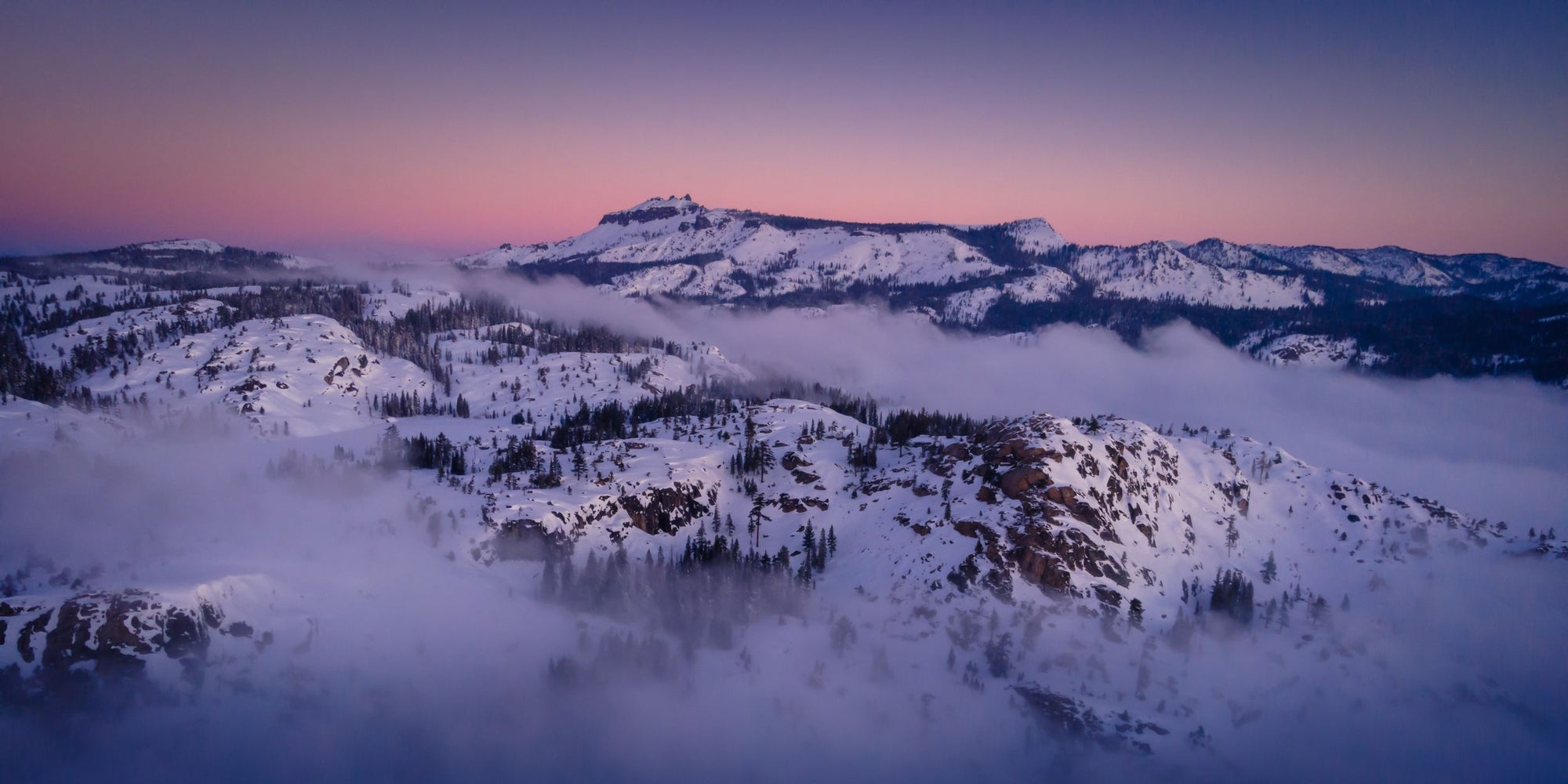 Fog lingers along snowcapped mountains that sit along a pink and purple horizon.