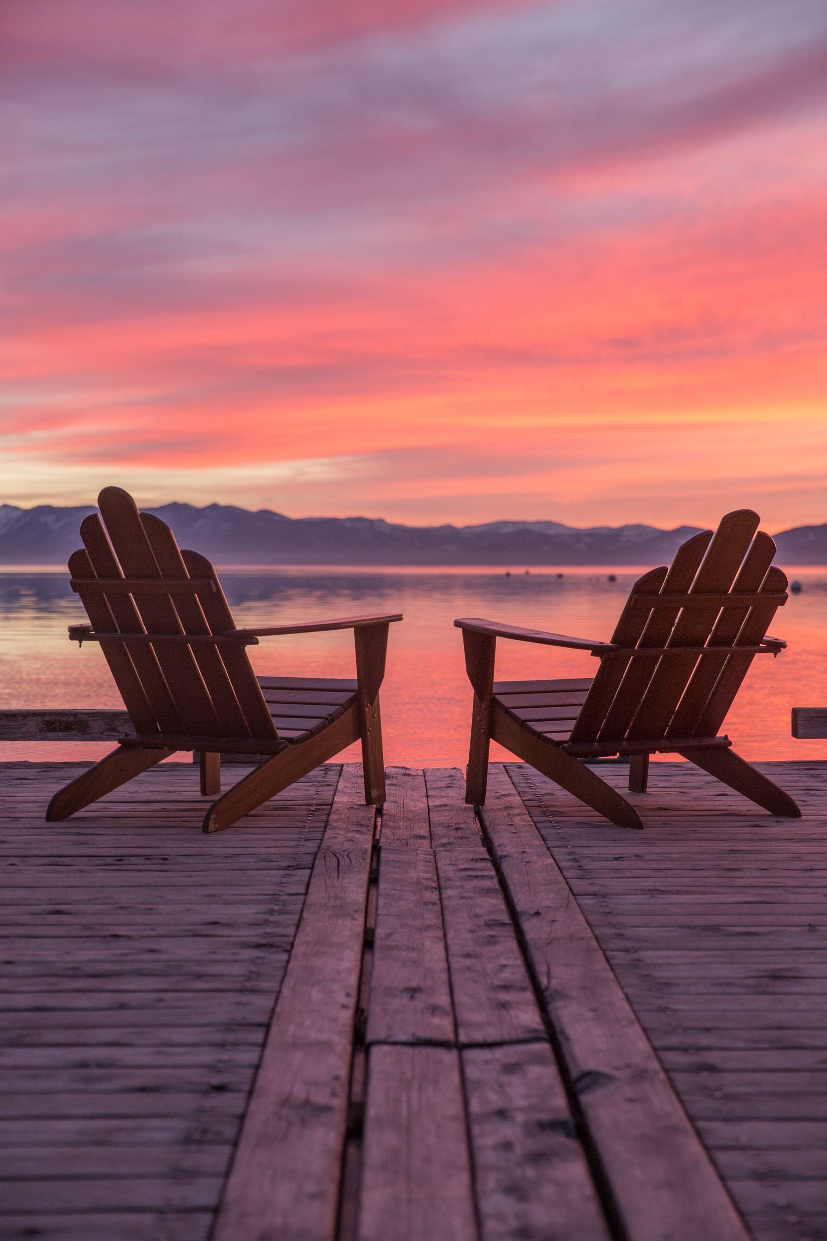 Two Adirondack chairs on a dock overlooking the a multi-color sunset above Lake Tahoe.