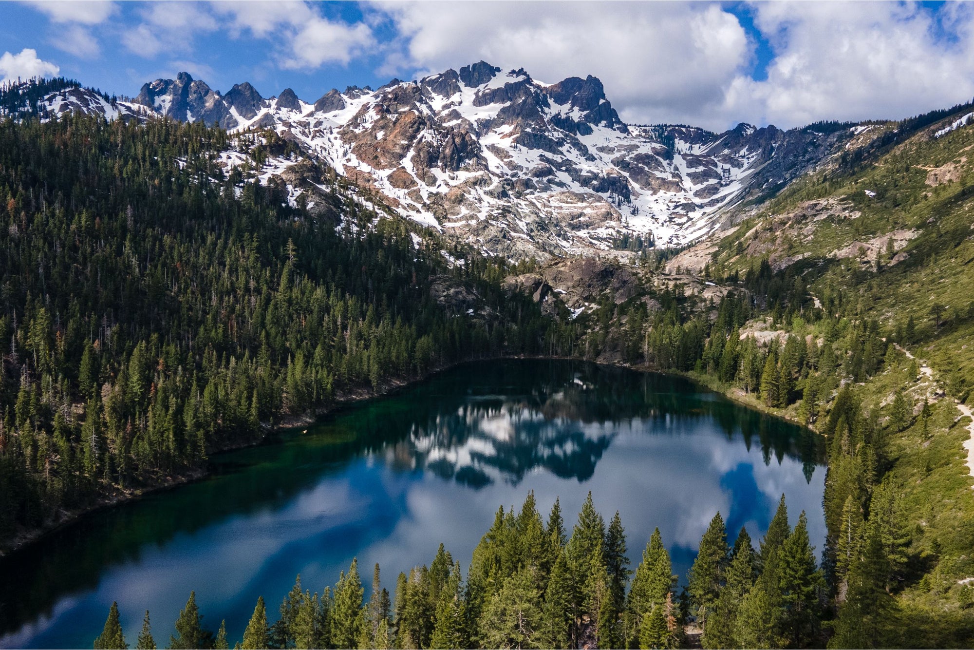 Mountain peaks capped with snow are reflected in an alpine lake lined with tall pine trees.