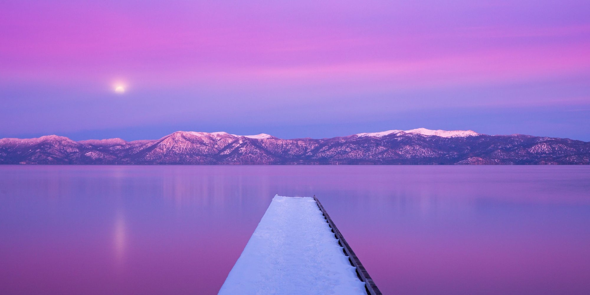 A snow covered dock juts out into Lake Tahoe as the sun sets and casts pink and purple hues across the landscape.