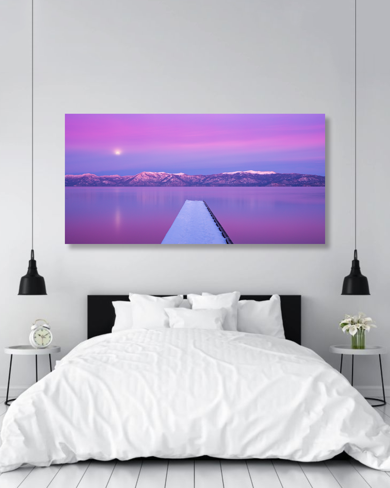 A large bed covered with a white duvet sits below a photo showcasing a pink and purple sunset over a dock on a lake.