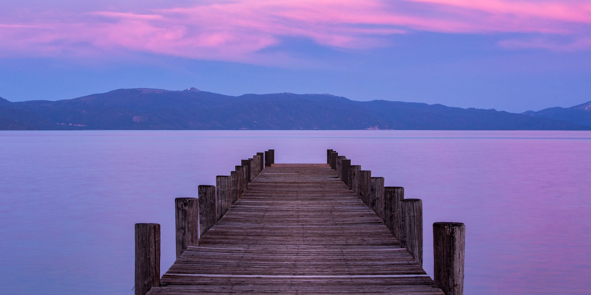 A wooden deck sits on Lake Tahoe as the horizon captures a pink and purple sunset.