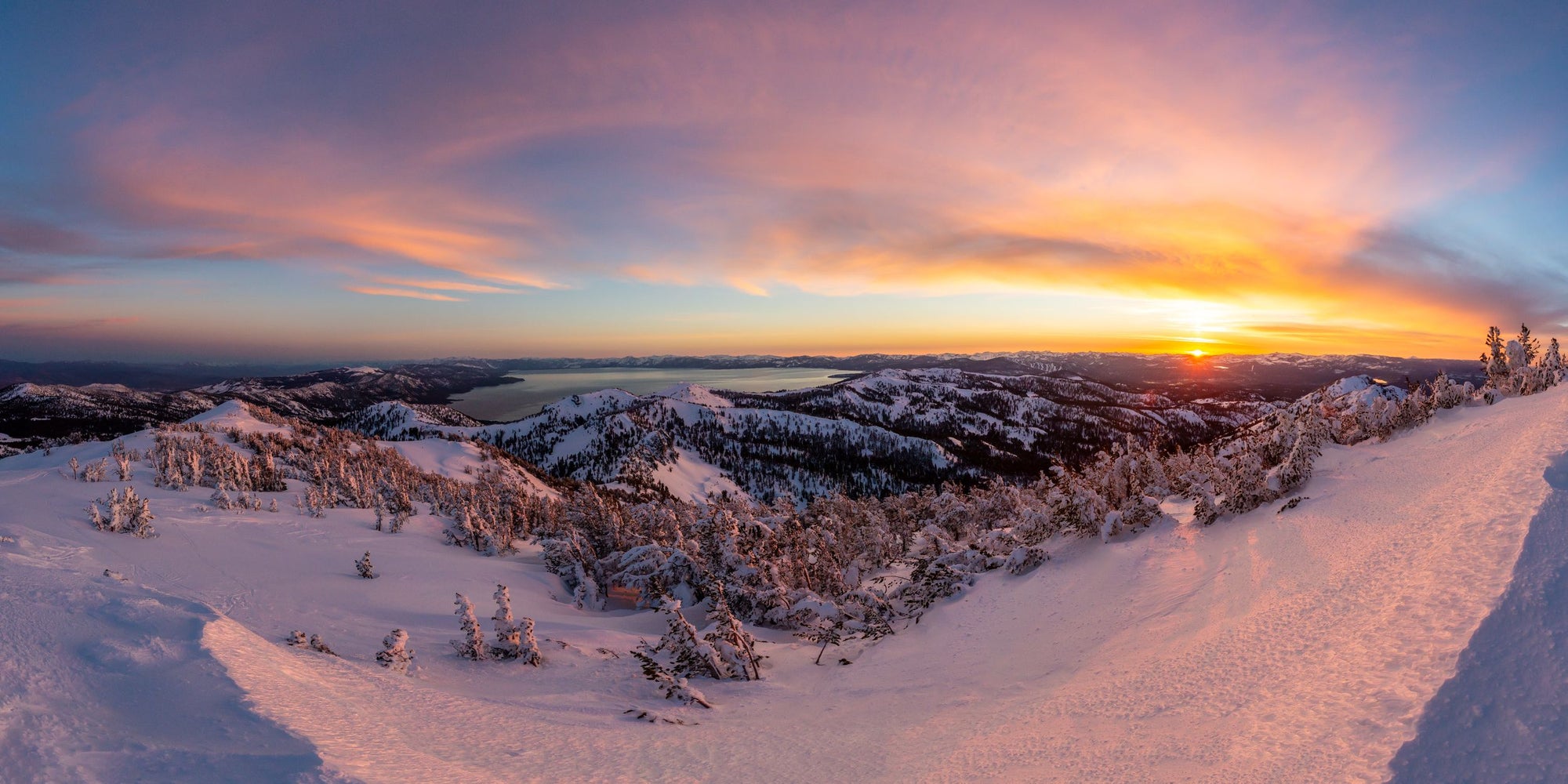 The sun rising above a lake surrounded by snowy mountains.